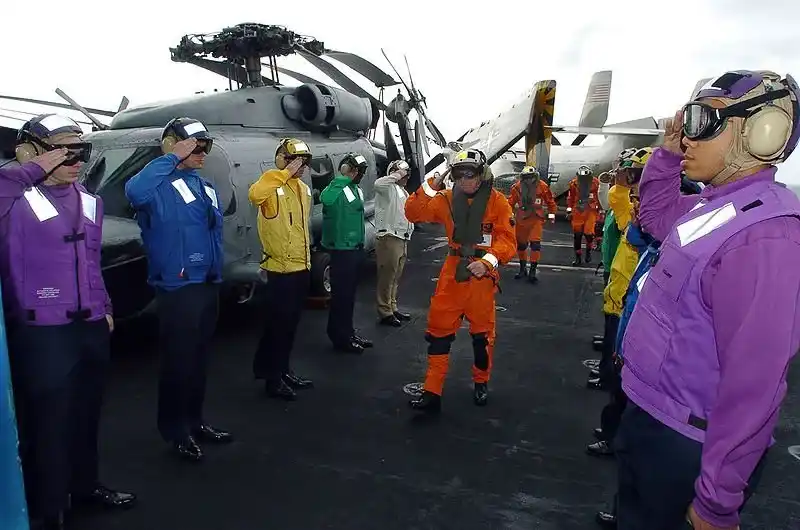 Aircraft carrier personnel in multi-colored jerseys coordinating launch under jet blast conditions